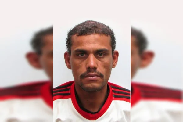 Imagem colorida de um homem negro, com cabelos curtos, cavanhaque e camiseta branca com detalhes em preto e vermelho na manga