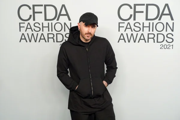A imagem mostra um homem vestido inteiramente de preto sob um fundo branco