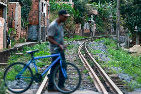 Imagem colorida mostra um homem negro conduzindo uma bicicleta sobre o trilhos de uma via férrea em TrÊs Rios, no Rio de janeiro