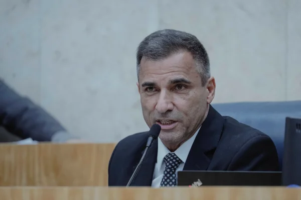 Imagem colorida mostra o vereador João Jorge, de terno e gravata, falando ao microfone