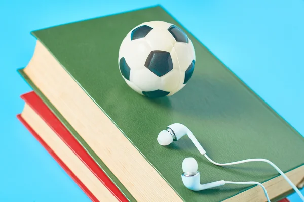 Fones de ouvido brancos, livros antigos, bola de futebol de brinquedo e bola de futebol americano sobre fundo azul. Conceito de audiolivro. Educação online. Aprendizagem a distância. Tecnologia moderna.