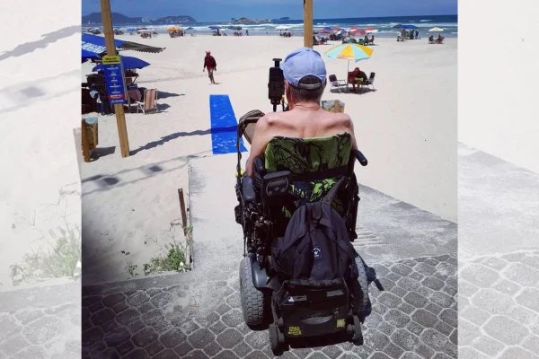 Imagem colorida mostra surfista Taiu Bueno em uma cadeira de rodas observando uma praia | Metrópoles