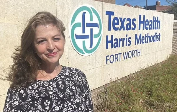 mulher de 61 anos curada do câncer no mesmo dia em que foi diagnosticada posa para foto na frente da placa do hospital onde foi operada