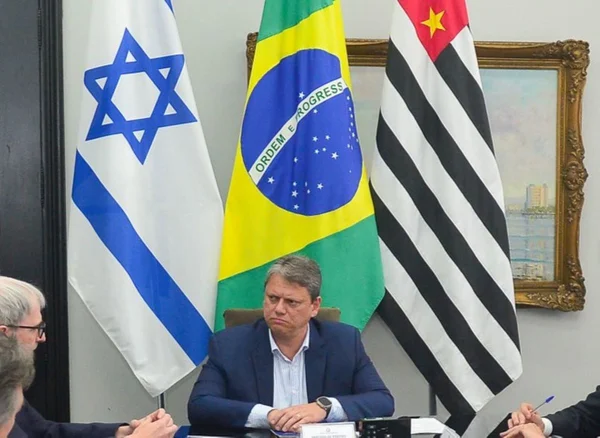 Tarcísio de Freitas sentado em uma mesa e ao fundo três bandeiras: uma de Israel, uma do Brasil e outra do Estado de SP - Metrópoles