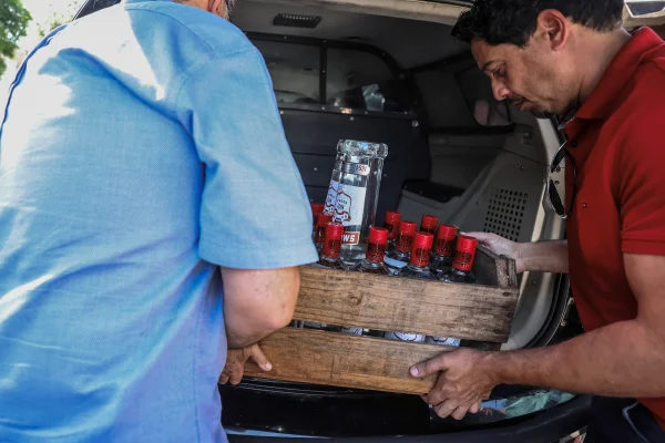 Imagem colorida de duas pessoas colocando caixa de vodca em porta-malas de carro. Metrópoles