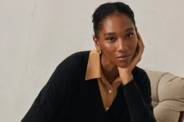 Na imagem com cor, modelo negra posa com mão no queixo enquanto veste cashmere preto com blusa marrom por baixo