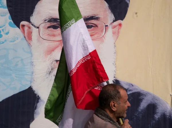 Imagem colorida de um cidadão do Irã carregando uma bandeira do país de frente a uma pintura do aiatolá Ali Khamenei - Metrópoles