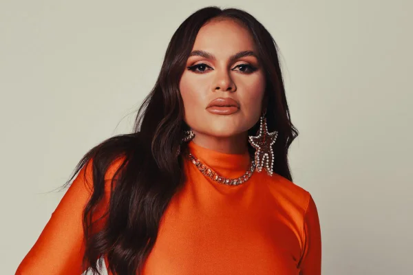 Foto colorida da cantora Samyra Show. Ela usa um vestido laranja e um brinco grande de estrela. Tem cabelo castanho e longo - Metrópoles