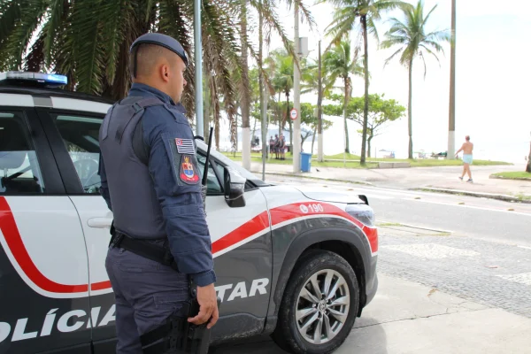Imagem colorida de policial militar ao lado de viatura perto da faixa de areia da praia. Metrópoles