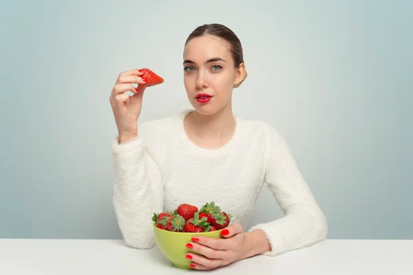 Foto colorida de mulher branca segurando um morango e uma tigela com a fruta - Metrópoles