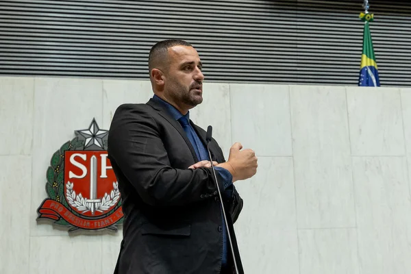 Deputado em tribunal da ALESP. Ele usa terno preto e, ao fundo, aparece o escudo da ALESP