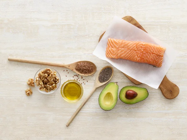 Foto mostra uma mesa com salmão, abacate, azeite de oliva, chia e nozes servidas, alimentos fontes de omega 3 e vitamina F