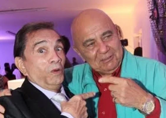 Dedé Santana de terno e Roberto Guilherme de camisa azul juntos apontando um para o outro