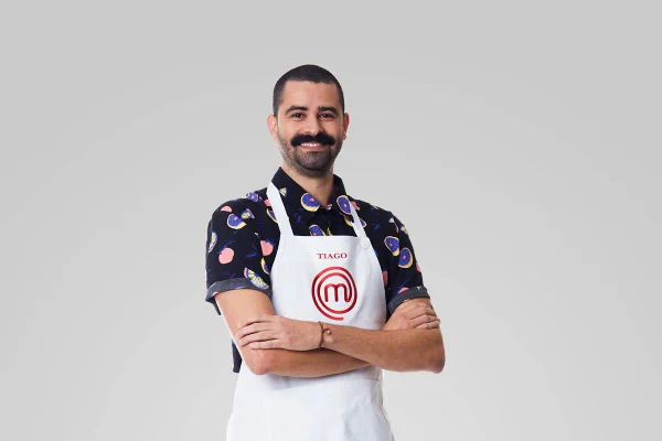 Homem com camisa de manga curta e avental do masterchef sorri com braços cruzados