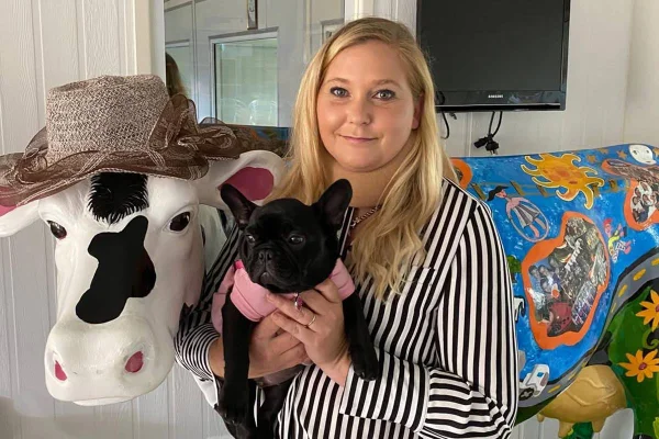 Foto colorida de mulher ao lado de escultura de vaca. Ela segura um cachorrinho - Metrópoles