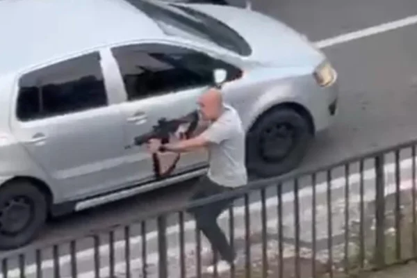 Homem de camiseta clara e calça jeans empunha fuzil em calçada de viaduto, em dia claro - Metrópoles