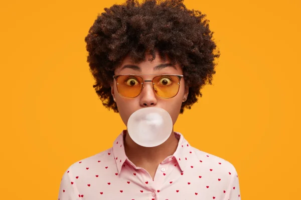 Foto colorida com fundo laranja de mulher fazendo bola com chiclete açucarado - Metrópoles