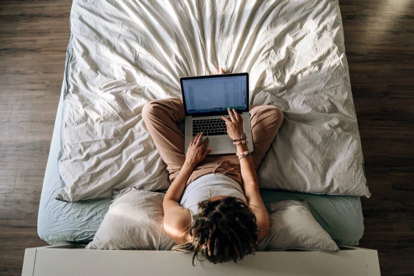 Foto colorida vista de cima de uma mulher com um computador no colo sentada na cama com lençol branco amassado - Metrópoles