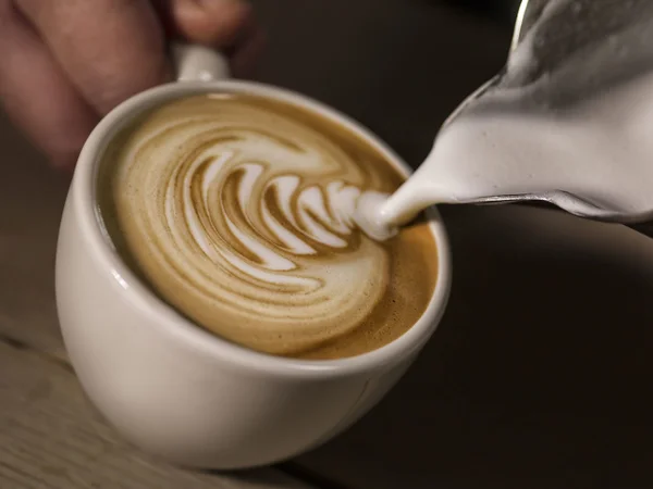 Na foto, uma mão segura uma xícara branca enquanto despeja leite vaporizado sobre o café - Metrópoles