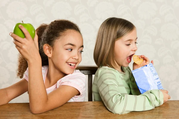 Criança comendo maçã e outra comendo batatinha