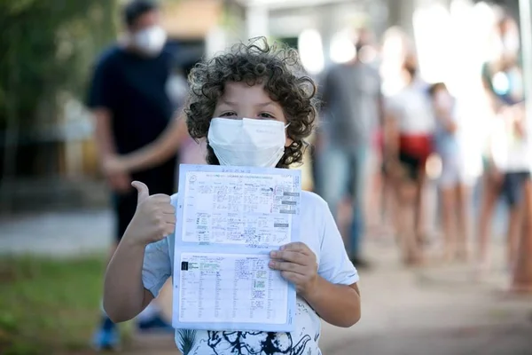 Menino de cabelo cacheado e máscara branca no rosto segurando papel e com a outra mão fechada e polegar para o alto