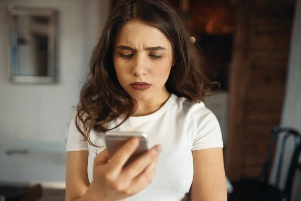 Close-up de uma jovem caucasiana séria e atraente segurando um smartphone, franzindo a testa ao ler uma notícia triste. Garota bonita usando o celular, digitando uma mensagem de texto, enfrentando problemas de conexão. Foco seletivo - Metrópoles