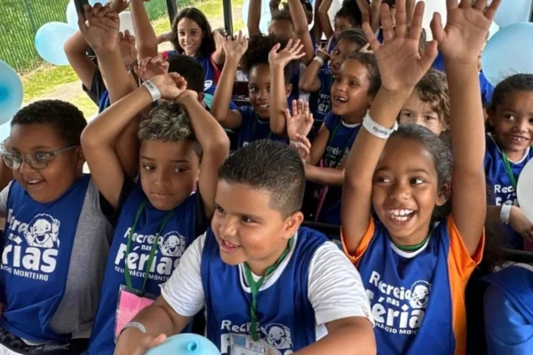 Imagem colorida mostra um grupo de crianças vestindo camiseta azul escrito Recreio nas Férias. Elas estão com expressão feliz e algumas têm os braços para cima - Metrópoles