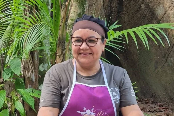 Imagem colorida mostra Dona Nena, uma mulher com uma touca no cabelo, camiseta e um avental roxo. Ela usa óculos e está sorrindo - Metrópoles