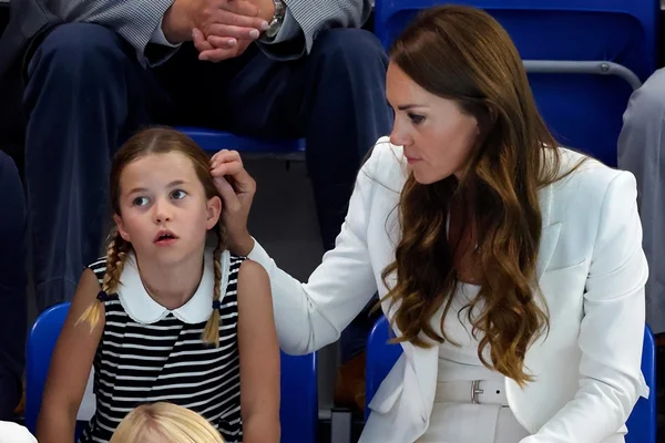 Charlotte está com um vestido listrado em preto e branco e usando tranças. Ao seu lado, Kate está olhando para a filha e passando a mão em seu cabelo. A princesa está usando um conjunto branco e cabelos soltos