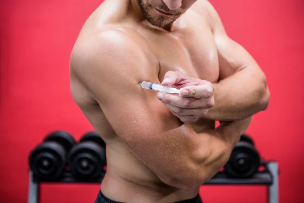 Foto colorida mostra homem branco injetando anabolizantes esterioides no braço em uma academia de paredes vermelhas - Metrópoles - Anvisa