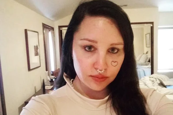 Foto colorida de Amanda Bynes. A atriz está com cabelo preto e liso, usa um moletom branco e está em casa. Ela tem um coração tatuado na bochecha - Metrópoles