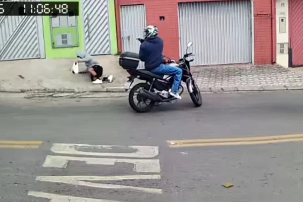 Imagem colorida de homem apontando arma para outro homem na calçada de uma rua. Metrópoles