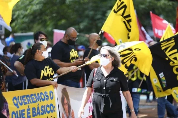 Pessoas estão segurando bandeiras e cartazes na cor amarela. Pessoas se aglomeram em um espaço público fazendo alguma espécie de manifestação e usam roupas pretas com as mesmas nomenclaturas com estampas. As blusas são pretas e as frases nelas têm a cor amarela