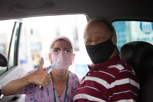 Mulher de máscara, roupa e touca rosa ao lado de um homem, dentro de um carro, posando para uma foto sorrindo e dando um joia com o dedão