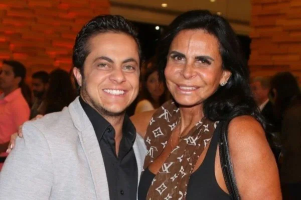 Foto colorida de homem branco com cabelos curtos e escuros, ele veste camisa presta e blazer claro; mulher branca de pele e cabelos morenos veste regata preta com cachecol marrom com logomarcas