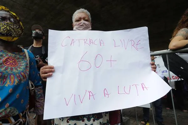 Fotografia colorida de mulher com cabelo grisalho segurando cartaz branco com escrita vermelha