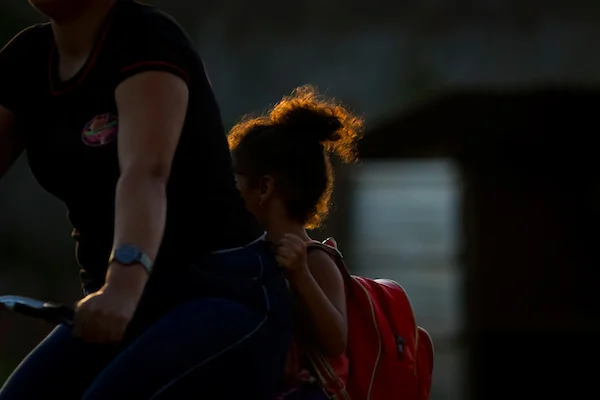 Menina com cabelo cacheado preso e mochila vermelha nas costas em garupa de mulher na bicicleta