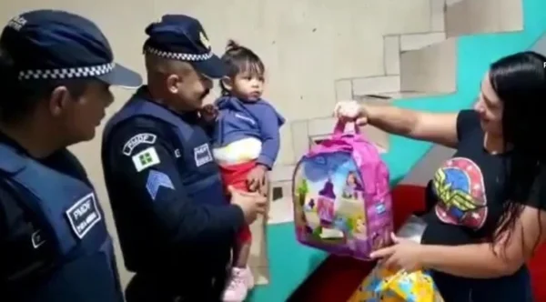 Policial militar segura criança que foi salva de engasgamento no colo e mãe mostra mochila recebida de presente pelos oficiais da corporação - Metrópoles
