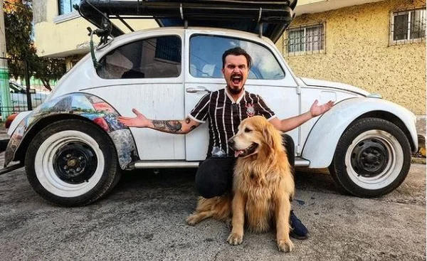 Foto colorida de um homem junto com um cachorro da raça golden retriver na frente de um fusca branco