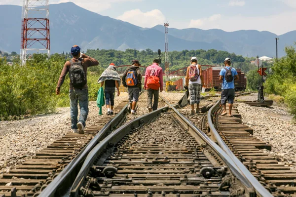 Foto colorida de imigrantes atravessando linha de trem - Metrópoles