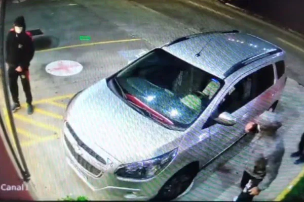 imagem colorida mostra cena filmada por câmera de segurança. três homens com moletons e máscaras estão ao redor de um carro de cor clara no estacionamento de uma farmácia - metrópoles