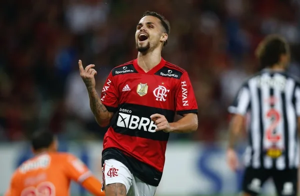 Na imagem colorida, um homem está posicionado no centro. Ele tem cabelos curtos, escuros e está usando uniforme de time esportivo