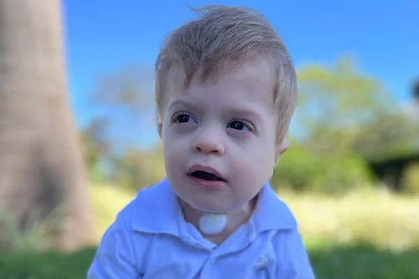 Foto colorida de menino loiro de blusa azul sentado em grama - Metrópoles