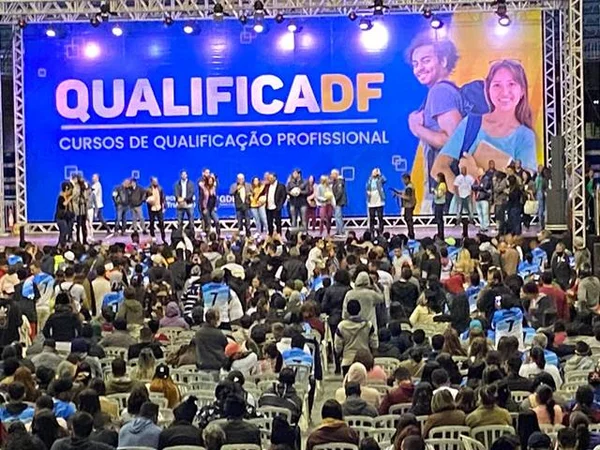 multidão de pessoas sentadas em um auditório. ao fundo um telão grande escrito qualifica DF