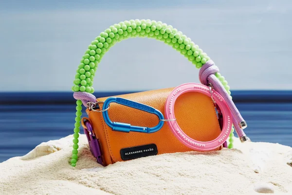 Bolsa modelo Olga da marca Alexandre Pavão. Ela é quadrada e de couro, na cor laranja, com uma alça verde e um mosquetão azul e outro rosa. - Metrópoles
