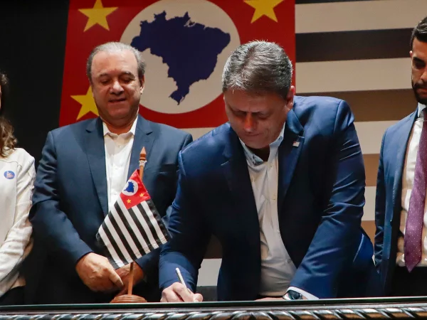 Imagem colorida mostra Tarcísio de Freitas em pé, apoiado em uma mesa enquanto assina um documento - Metrópoles