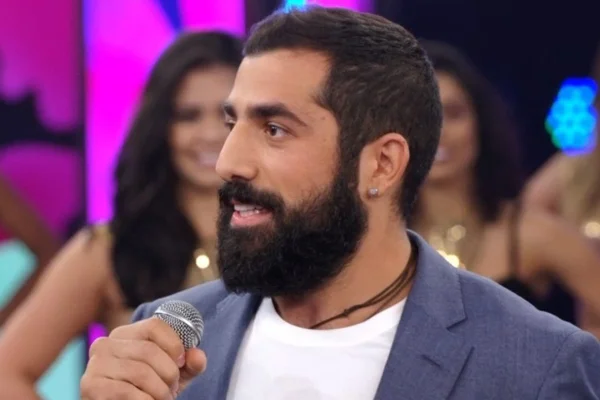 Foto colorida de Kaysar Dadour. Ele segura um microfone e usa um blazer azul com blusa branca - Metrópoles