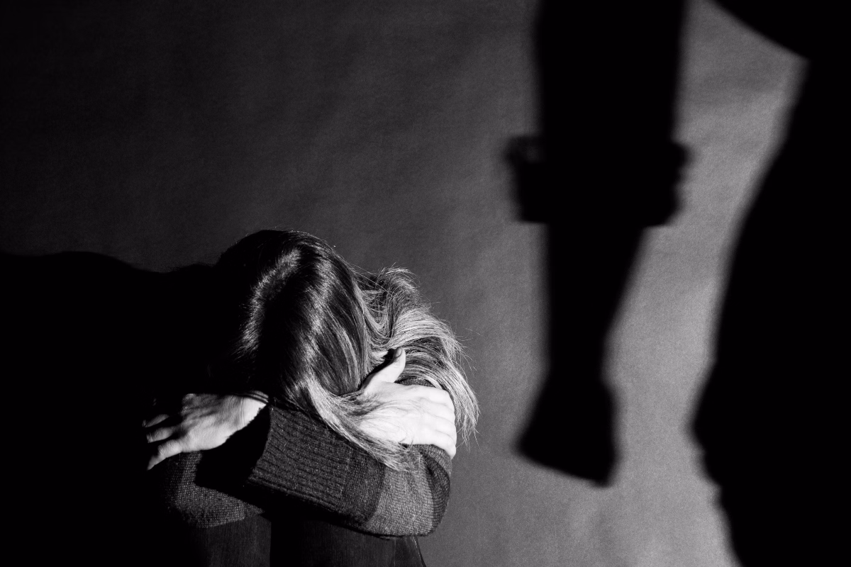 Imagem preto e branco de mulher acuada com cabeça apoiada nos joelhos, e sombra de mão masculina com punho fechado. Imagem conceito para violência contra mulher. - Metrópoles