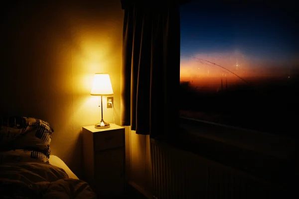 Candeeiro de cabeceira iluminando o canto do quarto ao lado da janela com vista para o céu ao entardecer