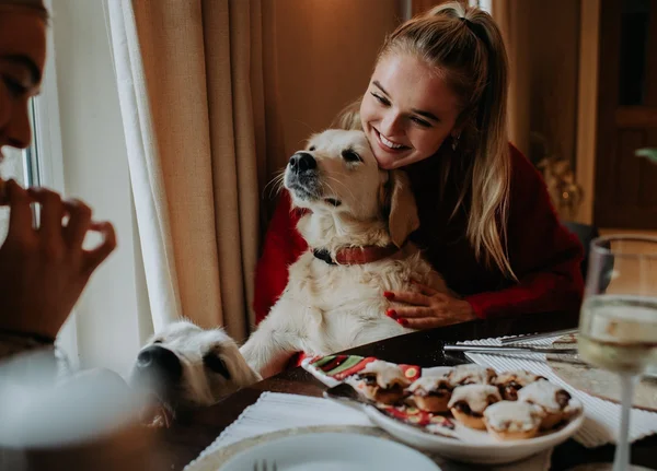 Mulher branca e jovem, de cabelo liso e loiro, faz carinho em um cachorro Golden. Eles estão na mesa durante a ceia de Natal. - Metrópoles